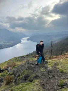 PawFecter dog walker Demi stood on the side of a hill with a dog with a large, long lake stretching into the distance